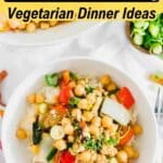 A bowl of vegetarian stir-fry with chickpeas, rice, and vegetables, topped with green onions, on a white table with additional dishes in the background.