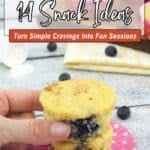 A hand holding two stacked blueberry muffins on a pink polka dot paper, with a tray of muffins and blueberries in the background. Text reads: "14 Snack Ideas - Turn Simple Cravings Into Fun Sessions.