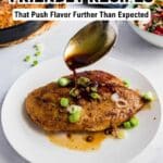 A spoon pours sauce over seasoned chicken breast garnished with chopped green onions on a white plate, with a side salad and skillet in the background.