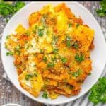 A plate of baked pasta with cheese and herbs is on a white plate, garnished with parsley, on a wooden table with a striped napkin and grated cheese nearby.