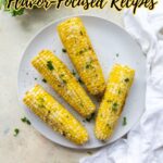 A white plate with four seasoned corn on the cobs, garnished with herbs, takes center stage on a light surface with fresh parsley nearby. Perfect for flavor-focused corn recipes. Text above and logo at the bottom.