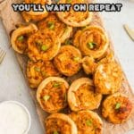A wooden board topped with several baked cheese pinwheels, garnished with green onions, with a small dish of dipping sauce beside them.