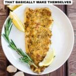 A baked fish fillet with a golden crust is served on a white plate with lemon wedges, rosemary sprigs, and garlic cloves.