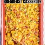 A baked tater tot breakfast casserole topped with chopped bacon, cheese, and green onions in a red dish, with a wooden spoon serving a portion.