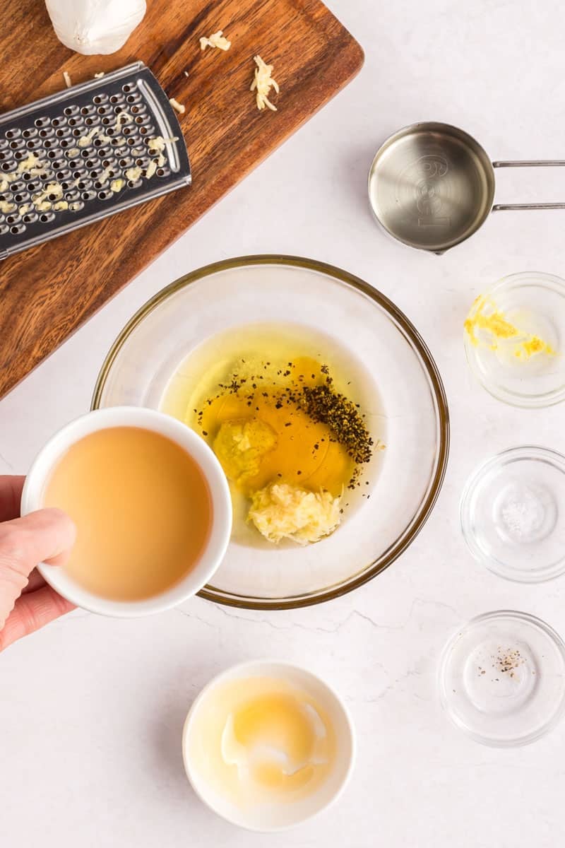Top-down view of a bowl with eggs, grated garlic, and spices being mixed, as a hand adds a small bowl of vinegar; other ingredients and utensils are arranged nearby.