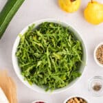 A bowl of arugula surrounded by a cucumber, two yellow pears, feta cheese, pomegranate seeds, walnuts, sunflower seeds, and small bowls of salt and pepper on a white surface.