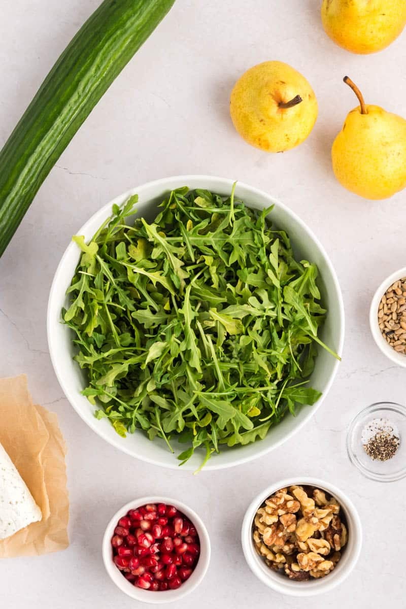 A bowl of arugula surrounded by a cucumber, two yellow pears, feta cheese, pomegranate seeds, walnuts, sunflower seeds, and small bowls of salt and pepper on a white surface.