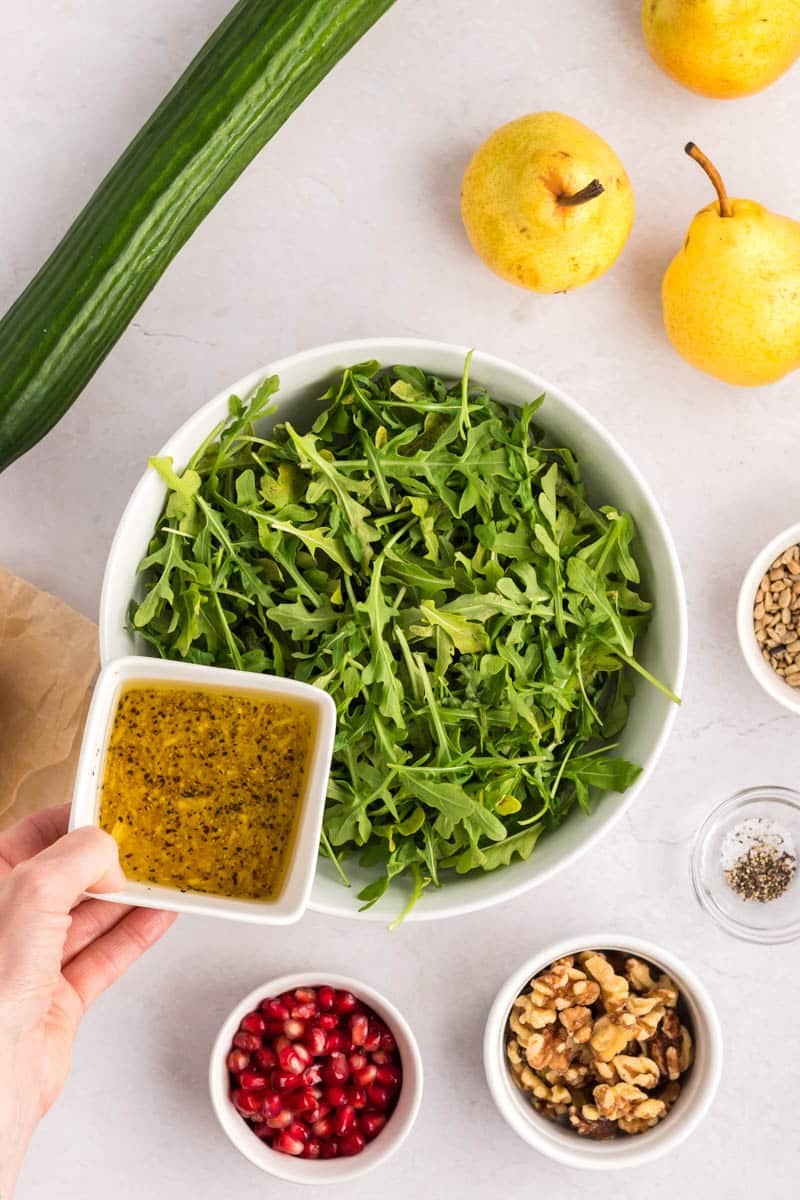 A bowl of arugula next to small bowls of pomegranate seeds, walnuts, sunflower seeds, pepper, a cucumber, two yellow pears, and a hand holding a bowl of dressing.
