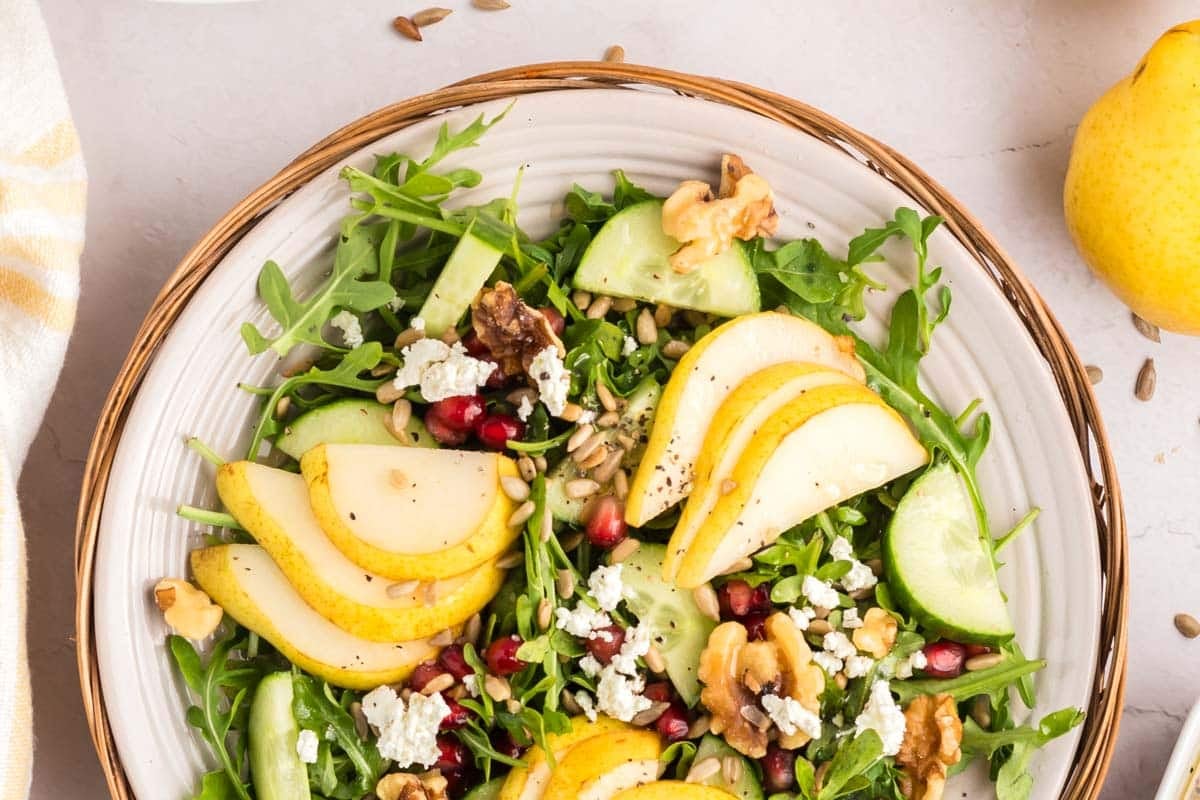 A salad with arugula, sliced pears, cucumber, walnuts, blue cheese, pomegranate seeds, and sunflower seeds on a white plate.