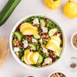 A bowl of salad with sliced pears, cucumber, walnuts, pomegranate seeds, arugula, and dollops of cheese, surrounded by ingredients on a white surface.