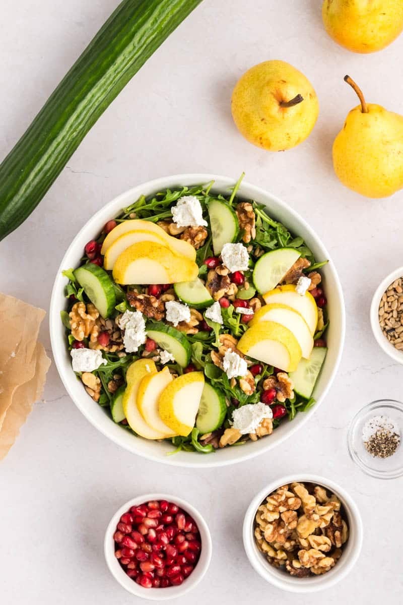 A bowl of salad with sliced pears, cucumber, walnuts, pomegranate seeds, arugula, and dollops of cheese, surrounded by ingredients on a white surface.