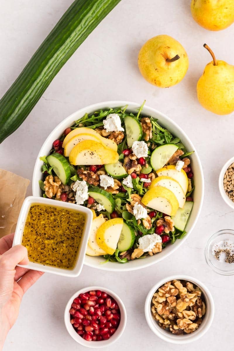 A hand holds a bowl of salad dressing over a salad with arugula, sliced pears, cucumbers, walnuts, goat cheese, and pomegranate seeds. Ingredients are arranged around the bowl.