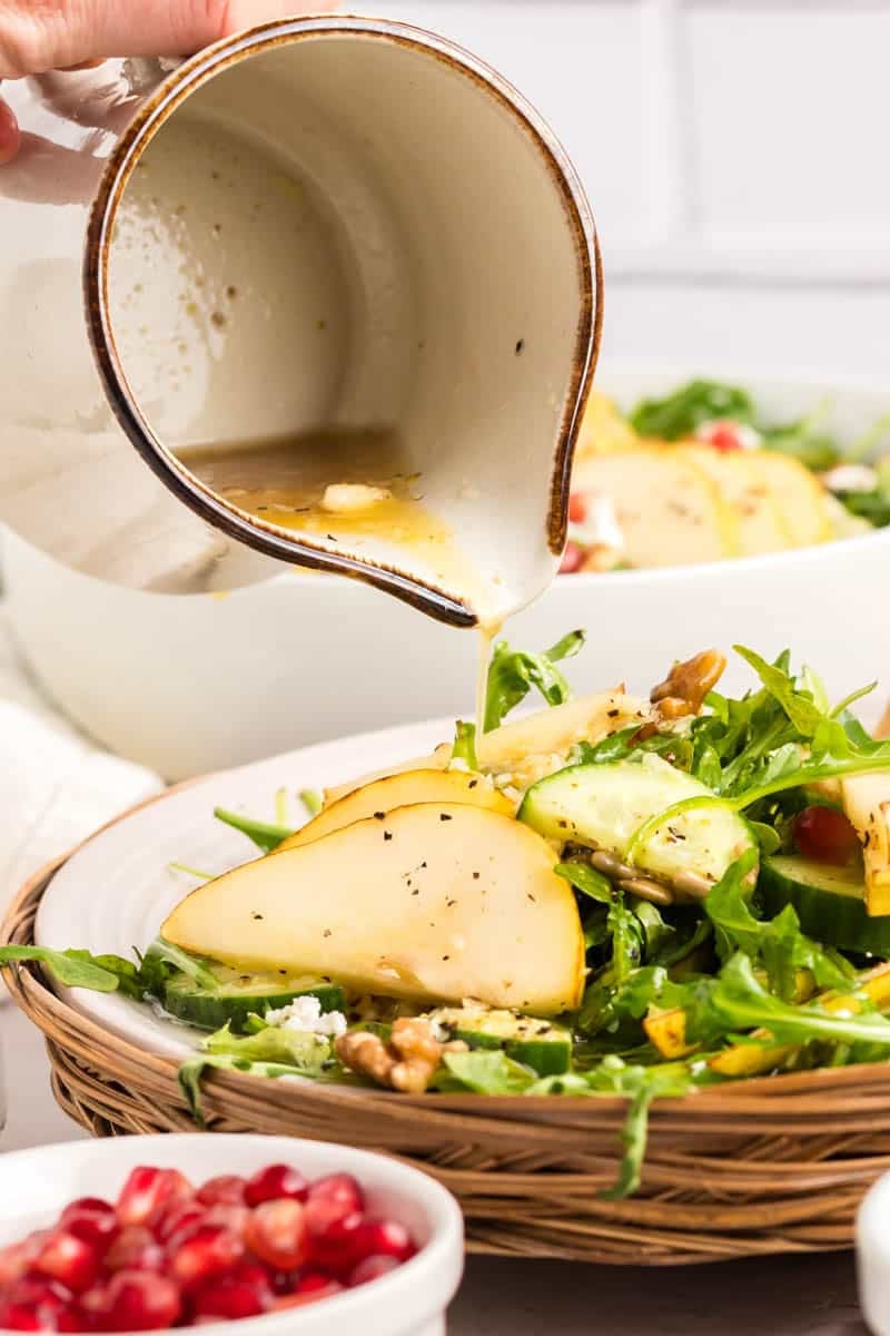 A hand pours dressing from a white ceramic pitcher onto a salad with arugula, pear slices, walnuts, and cheese on a plate. A bowl of pomegranate seeds is in the foreground.