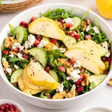 A bowl of salad with sliced pears, cucumbers, arugula, walnuts, pomegranate seeds, crumbled cheese, and black pepper.