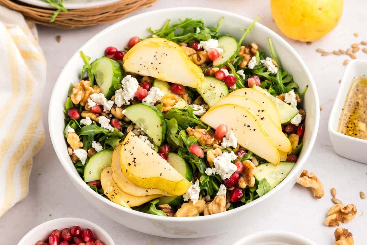 A bowl of salad with sliced pears, cucumbers, arugula, walnuts, pomegranate seeds, crumbled cheese, and black pepper.