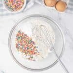 A glass bowl with flour and rainbow sprinkles, a whisk inside, next to two eggs in a tray and a small bowl of sprinkles on a marble surface.
