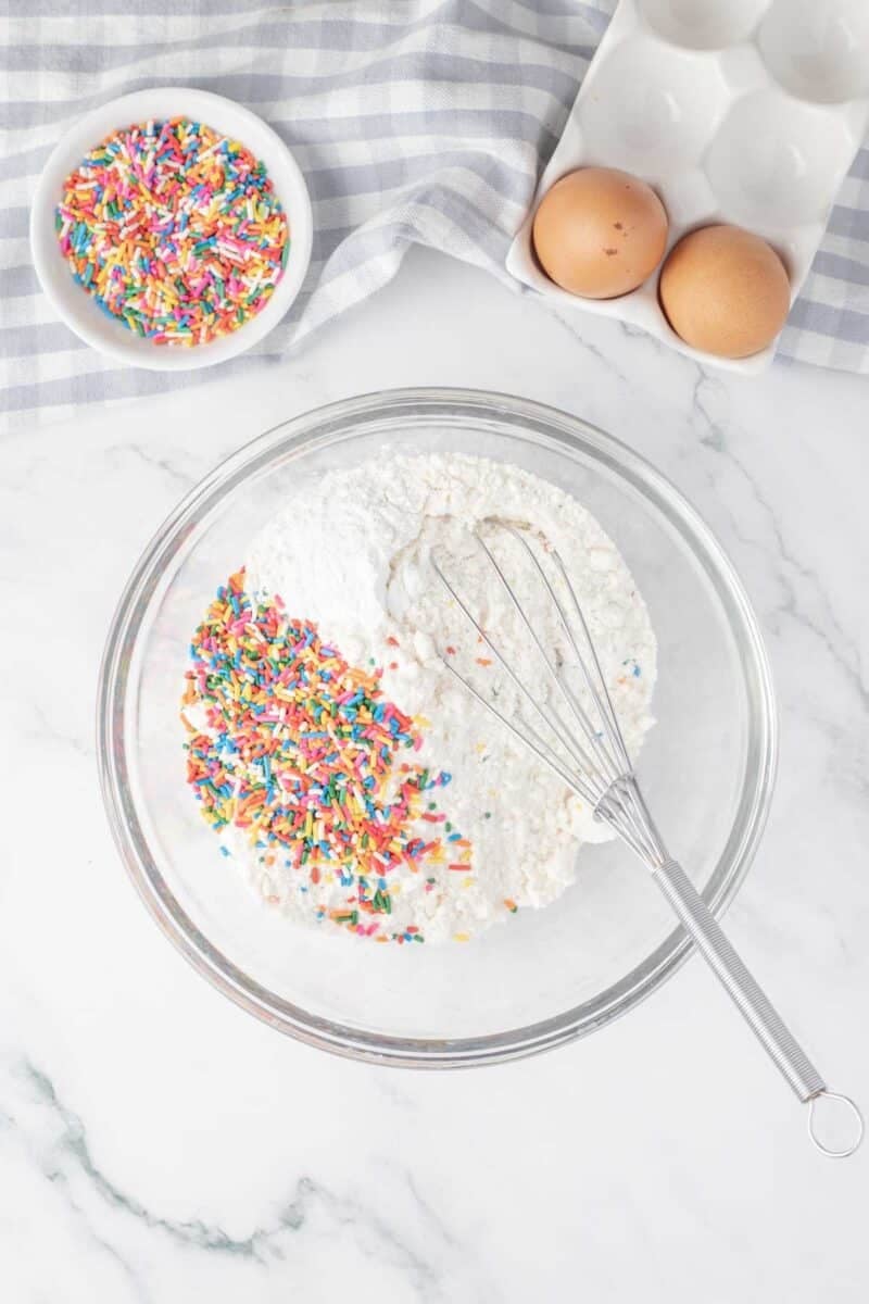 A glass bowl with flour and rainbow sprinkles, a whisk inside, next to two eggs in a tray and a small bowl of sprinkles on a marble surface.