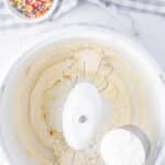 A mixing bowl with creamed ingredients, a beater attachment, and a half-cup of powdered sugar being added; a bowl of sprinkles and a checkered cloth in the background.