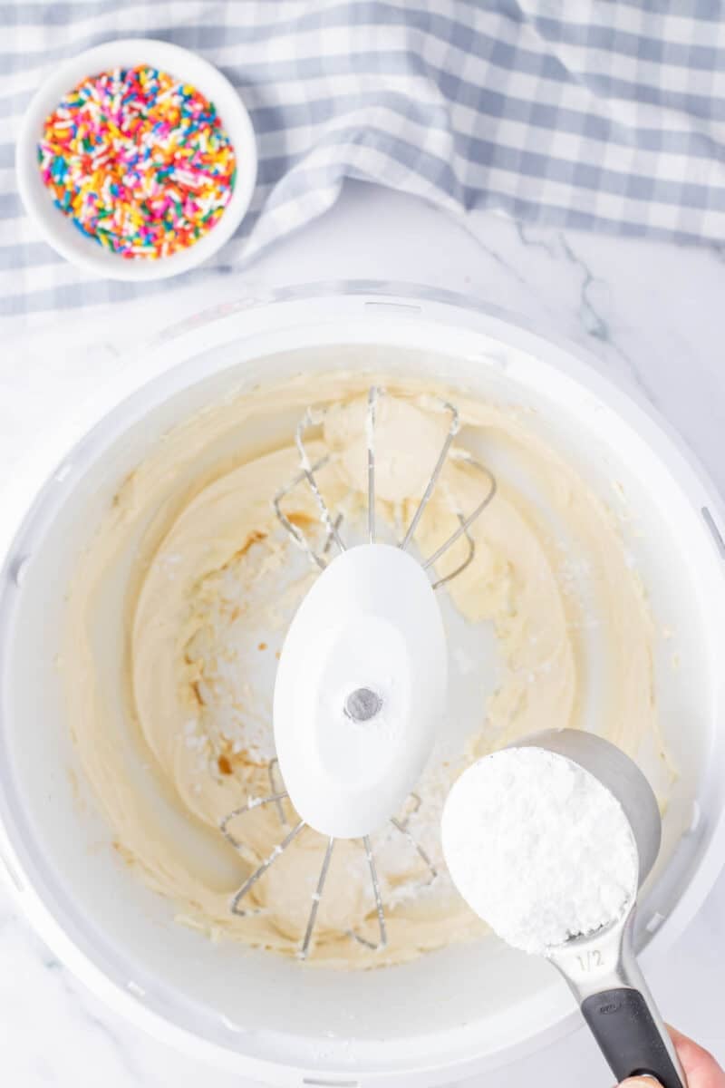 A mixing bowl with creamed ingredients, a beater attachment, and a half-cup of powdered sugar being added; a bowl of sprinkles and a checkered cloth in the background.