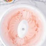 A mixing bowl with pink frosting and beaters, next to a small bowl of rainbow sprinkles on a marble countertop with a checkered cloth.
