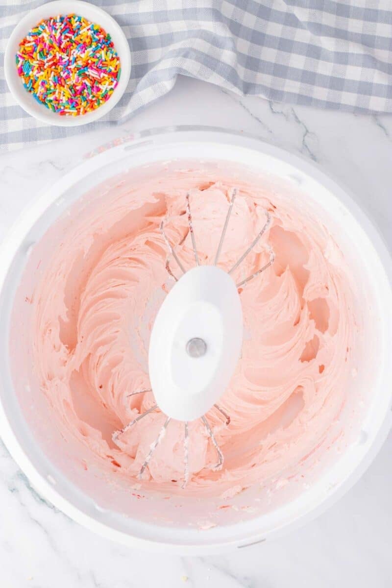 A mixing bowl with pink frosting and beaters, next to a small bowl of rainbow sprinkles on a marble countertop with a checkered cloth.