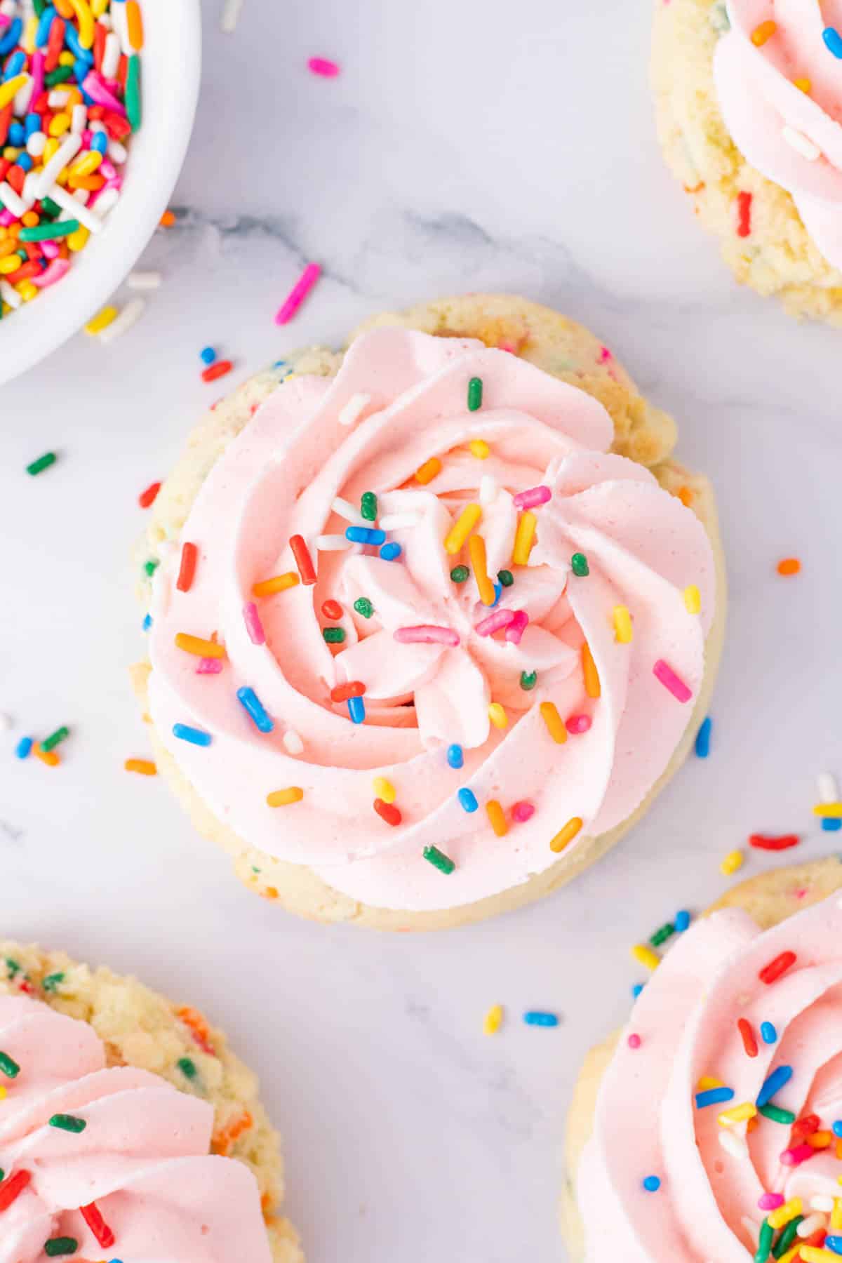 A birthday cake cookie topped with pink swirled frosting and colorful rainbow sprinkles on a white surface.