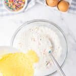 A glass bowl with dry cake mix and sprinkles, a bowl of yellow batter being poured in, two eggs, and a bowl of sprinkles on a marble counter.
