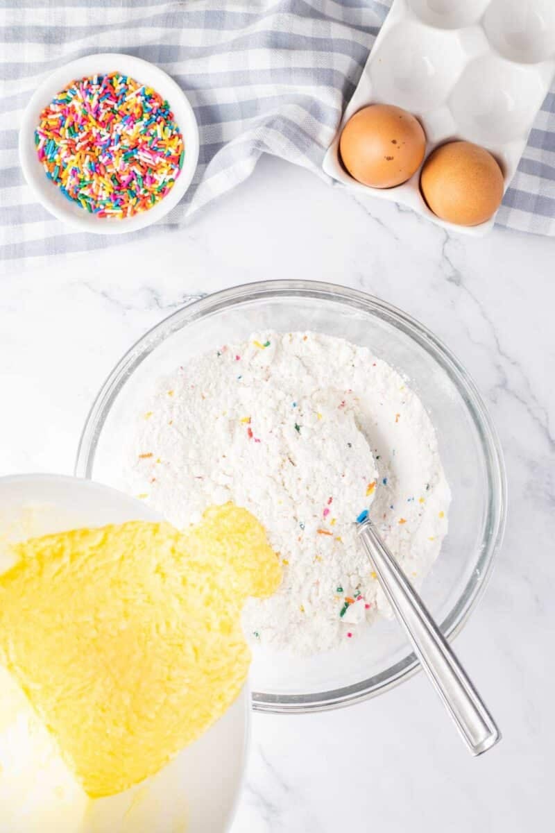 A glass bowl with dry cake mix and sprinkles, a bowl of yellow batter being poured in, two eggs, and a bowl of sprinkles on a marble counter.