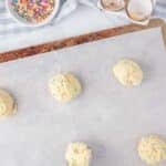 Five scoops of cookie dough with sprinkles on a parchment-lined baking sheet, with a bowl of sprinkles, broken eggshells, and a kitchen towel nearby.