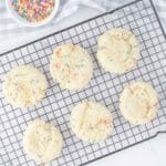 Six round sugar cookies with colorful sprinkles are cooling on a wire rack. A small bowl of sprinkles is set nearby on a checkered cloth.