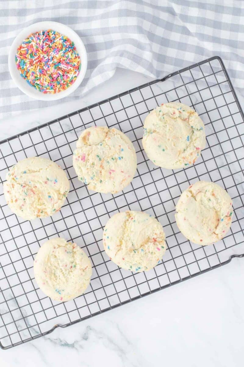 Six round sugar cookies with colorful sprinkles are cooling on a wire rack. A small bowl of sprinkles is set nearby on a checkered cloth.