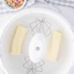 Two sticks of butter in a stand mixer bowl with paddle attachments, next to a small bowl of colorful sprinkles on a white surface with a checkered cloth.