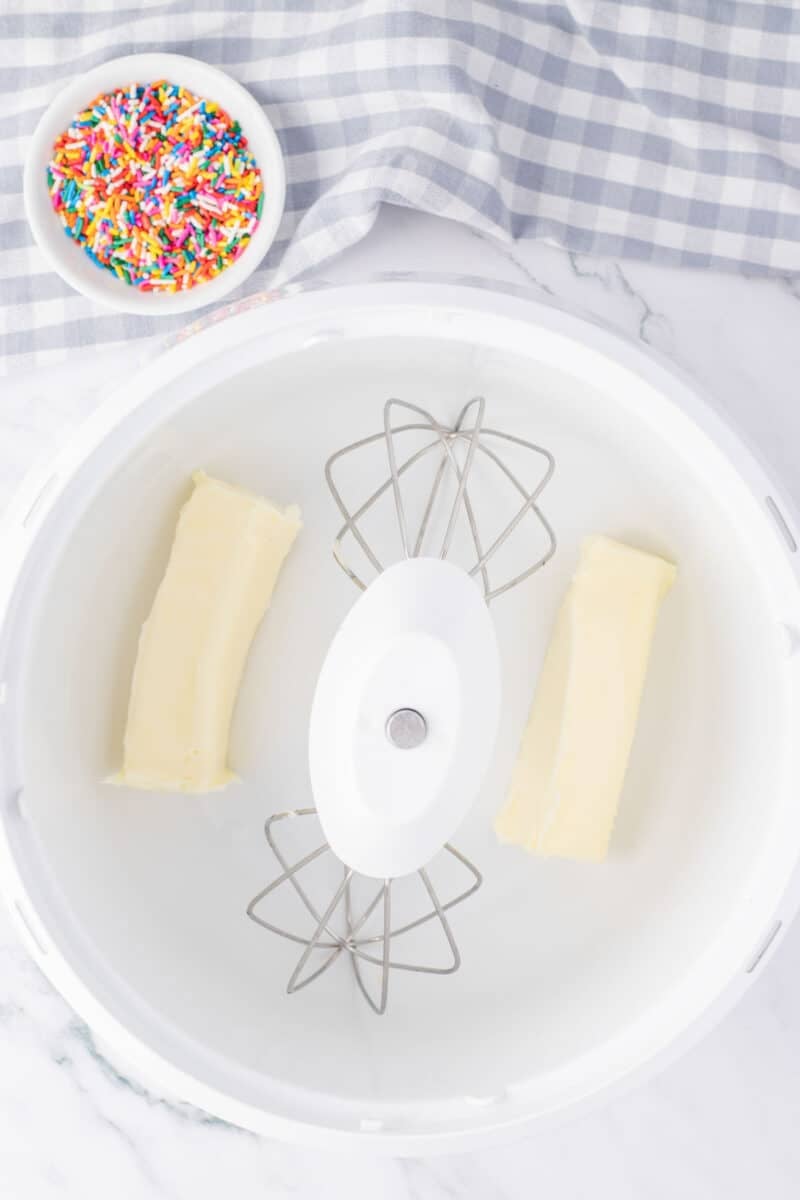 Two sticks of butter in a stand mixer bowl with paddle attachments, next to a small bowl of colorful sprinkles on a white surface with a checkered cloth.