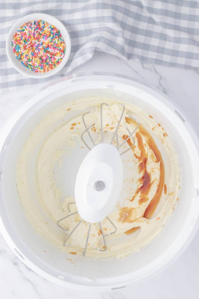 A stand mixer bowl with partially mixed batter and vanilla extract, next to a small bowl of colorful sprinkles on a marble countertop.