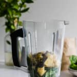 A blender pitcher containing spinach, pineapple chunks, banana pieces, and chia seeds sits on a white countertop.