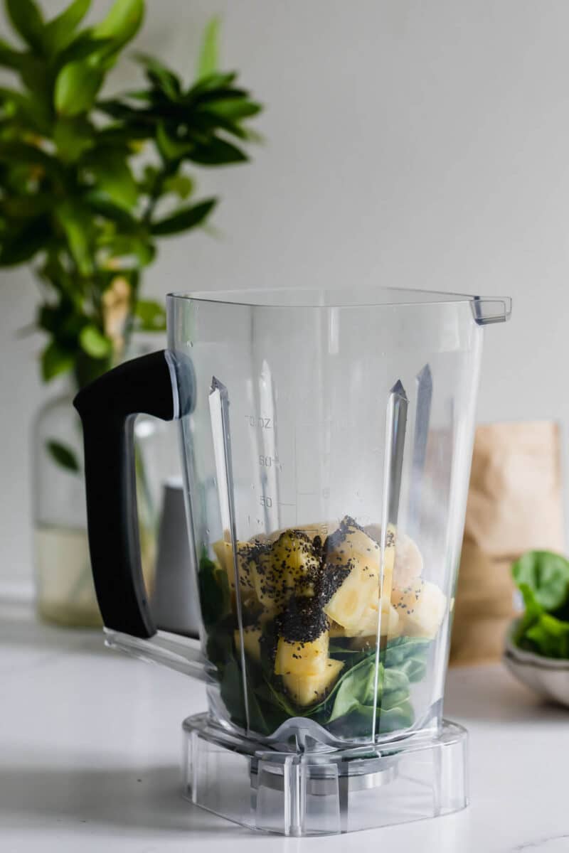 A blender pitcher containing spinach, pineapple chunks, banana pieces, and chia seeds sits on a white countertop.