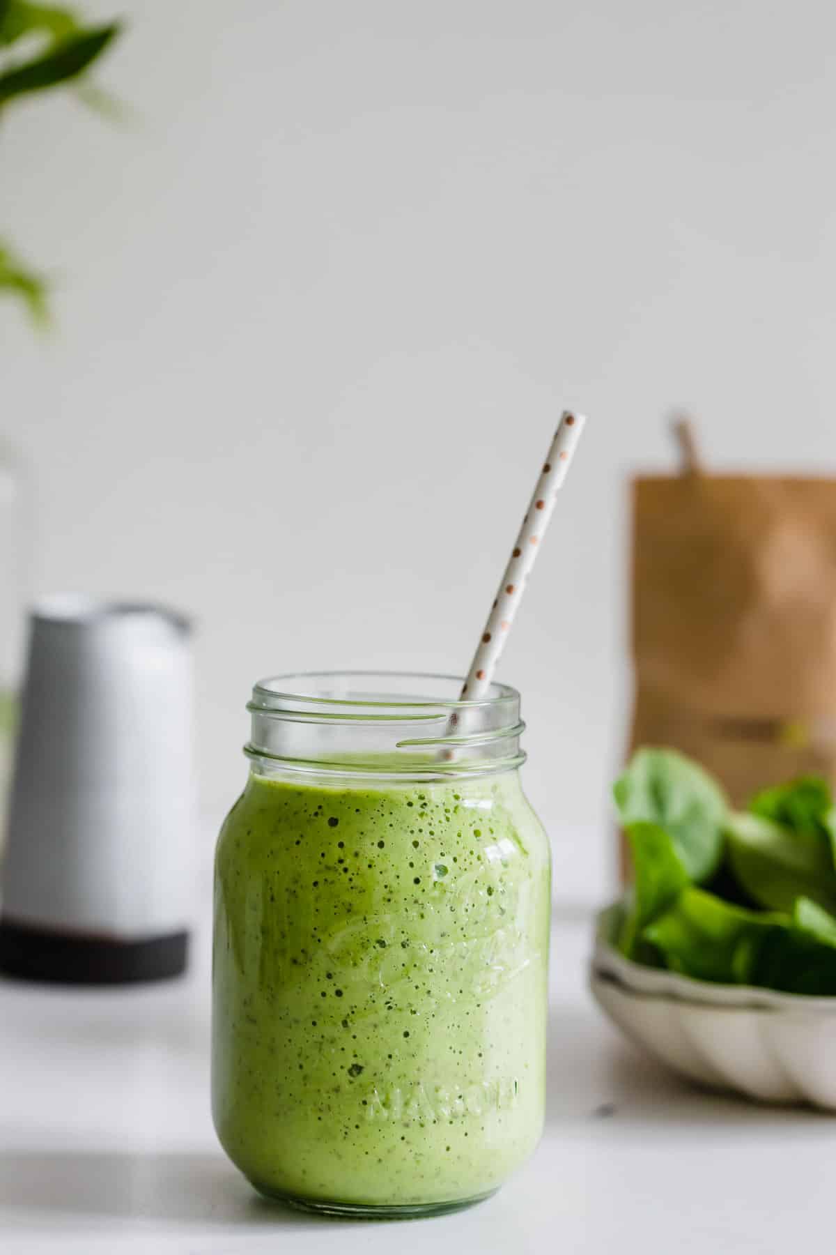 A green breakfast smoothie in a mason jar with a spotted straw sits on a white surface, with spinach leaves and a brown paper bag in the background.