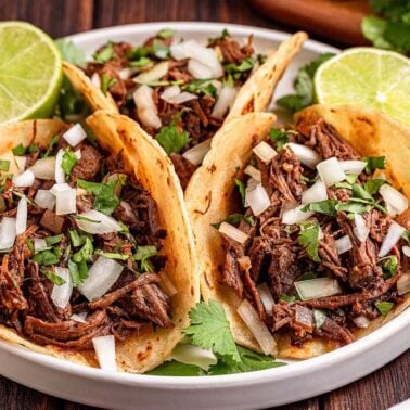 Three beef tacos topped with chopped onions and cilantro, served on a plate with lime halves and garnished with fresh cilantro—a perfect choice for flavorful dinners.