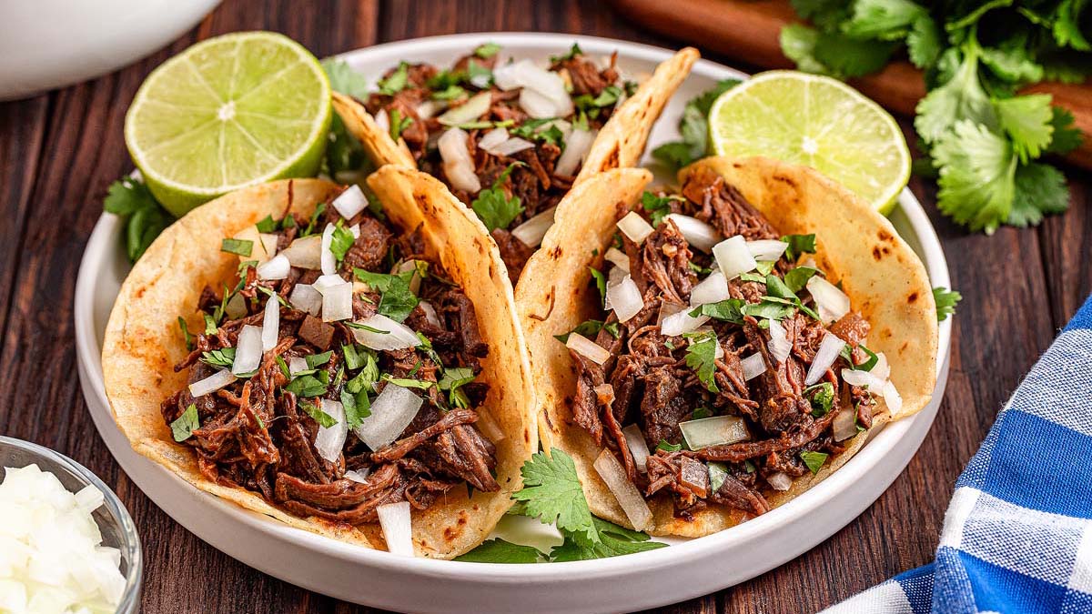 Three beef tacos topped with chopped onions and cilantro, served on a plate with lime halves and garnished with fresh cilantro—a perfect choice for flavorful dinners.
