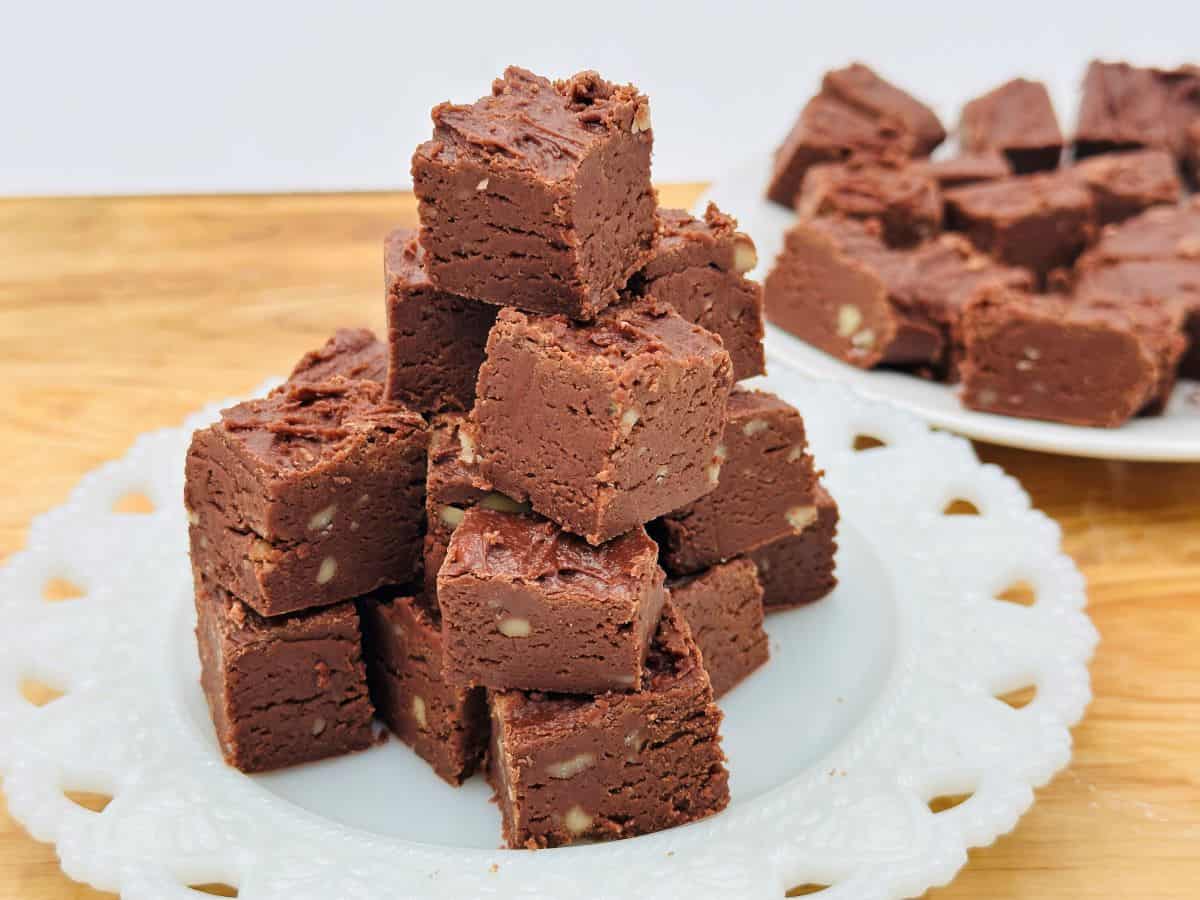 A plate stacked with square pieces of chocolate fudge with visible nut pieces, set on a wooden surface—a tempting treat for any sweet tooth; more fudge pieces are on a plate in the background, perfect inspiration for fudge recipes.