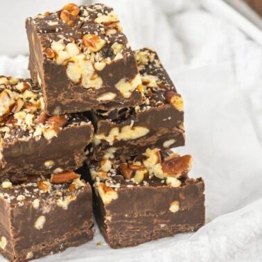 Five pieces of rich chocolate fudge with chopped nuts on top, stacked on white parchment paperโperfect for anyone with a sweet toothโwith more tempting fudge pieces in the background.
