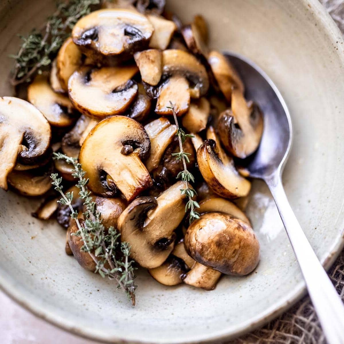 A bowl of sautéed sliced mushrooms, rich in umami flavor, is garnished with sprigs of fresh thyme and served with a metal spoon on the side.