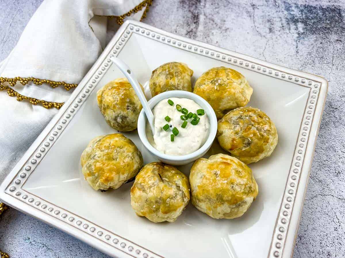 Seven pastry-wrapped beef appetizers are arranged in a circle on a square plate around a small bowl of creamy dip topped with chopped chives. A white napkin is placed nearby, making this one of the perfect party trays for any gathering.