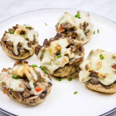 A white plate with five stuffed mushrooms topped with melted cheese and garnished with chopped green herbsโperfect beef appetizers for party trays.