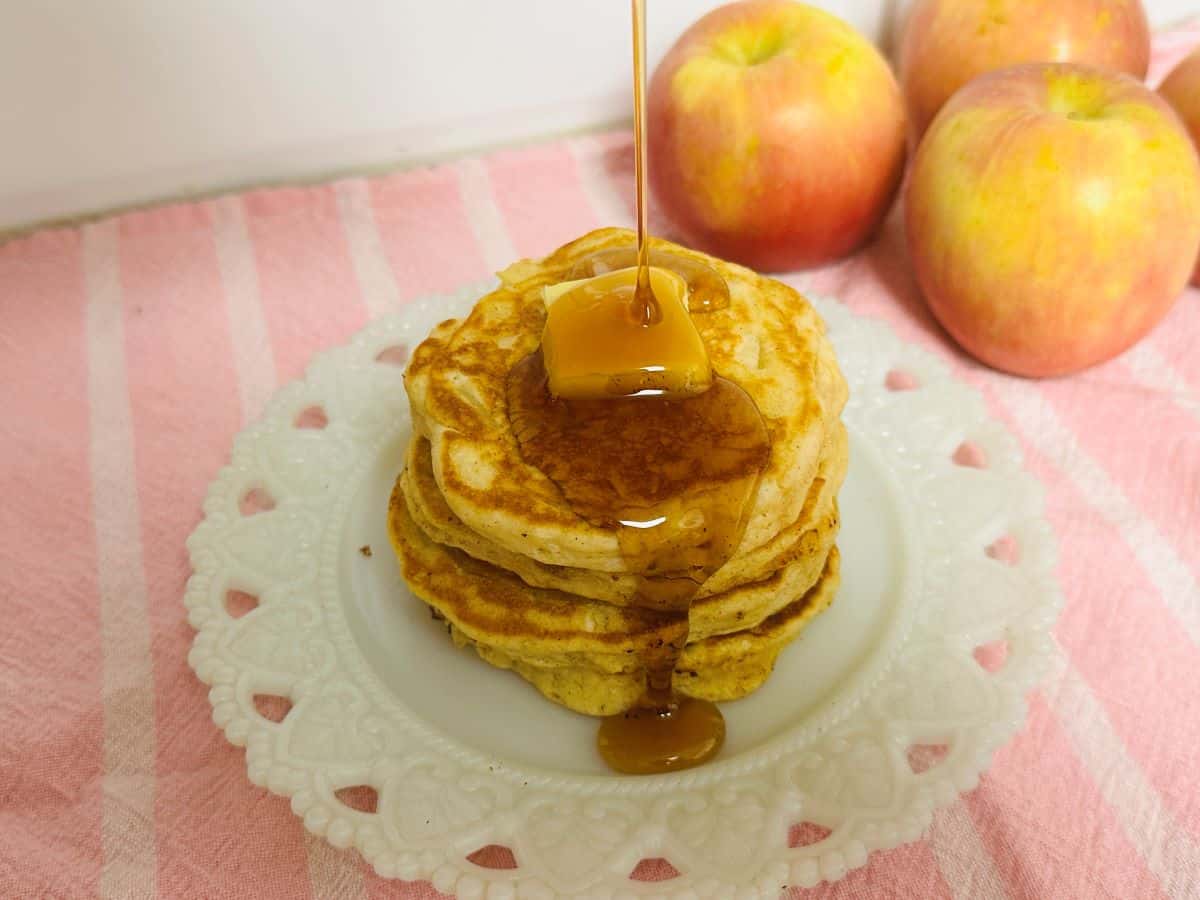 A stack of fluffy pancakes topped with a pat of butter and syrup being poured, with three apples in the background on a pink striped cloth—perfect for a delicious breakfast.