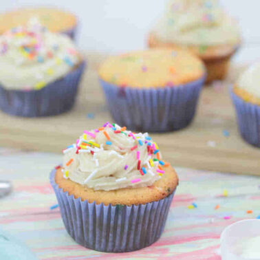 Vanilla cupcakes with a subtle flavor, white frosting, and colorful sprinkles in purple wrappers are arranged on a wooden board and pastel surfaceโperfect for inspiring new vanilla ideas.