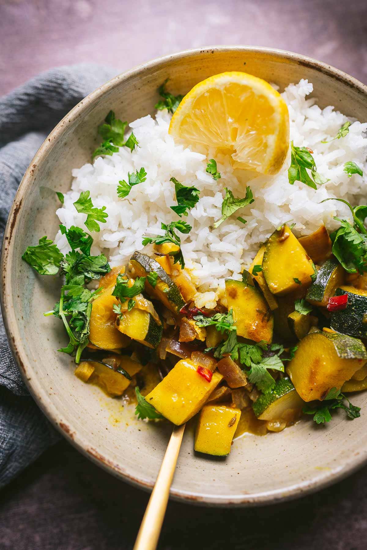 A bowl of white rice topped with chopped cilantro, served alongside a comforting curry with zucchini and other vegetables, garnished with a lemon slice.