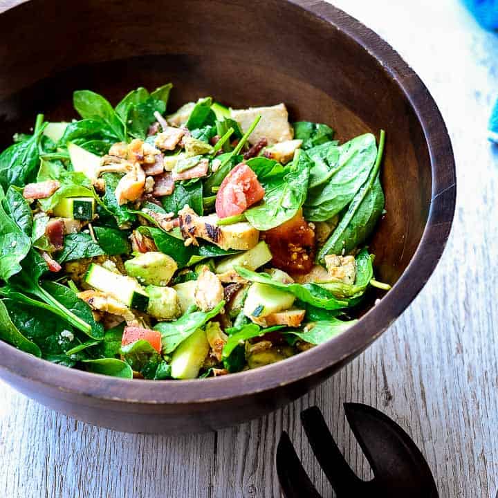 A wooden bowl filled with spinach salad, diced tomatoes, cucumbers, avocado, and bacon pieces offers a smoky flavor, placed on a light wooden surface with wooden salad utensils nearby.