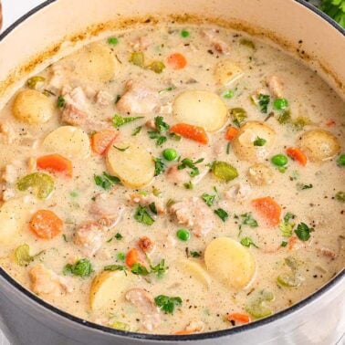 A pot of creamy chicken stew with potatoes, carrots, celery, peas, and herbs sits on a white surface next to bread, parsley, and salt and pepper shakers, radiating slow warmth and cozy flavorโa true classic among stew recipes.