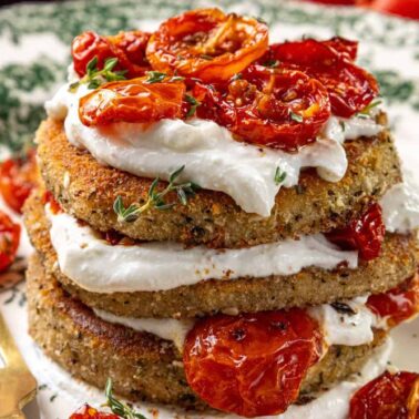 A stack of breaded fried eggplant slices layered with ricotta for a creamy texture and roasted cherry tomatoes on a decorative plate, with a fork and knife beside it.